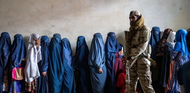 A man with a gun stand next to a long line of women who are covered from head to toe in a hijab.