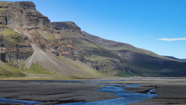 A photo of one side of a mountain with a clear cyan sky above. The foreground is a dark-coloured gravel bed with small streams trickling through it. The water is reflecting the blue of the sky. The mountainside has large patches of green vegetation mixed with scree slopes and layered palagonite. Some of the bluffs have flat faces and tops, and the peaks run diagonally from top left to below centre right. A small white horsetail waterfall is dropping near the lower centre of the shot, reaching ground level behind a scree pile.