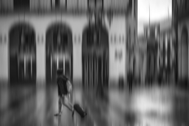 Fotografía en blanco y negro y con desenfoque de movimiento donde se ve una figura con maleta en unas calles mojadas.