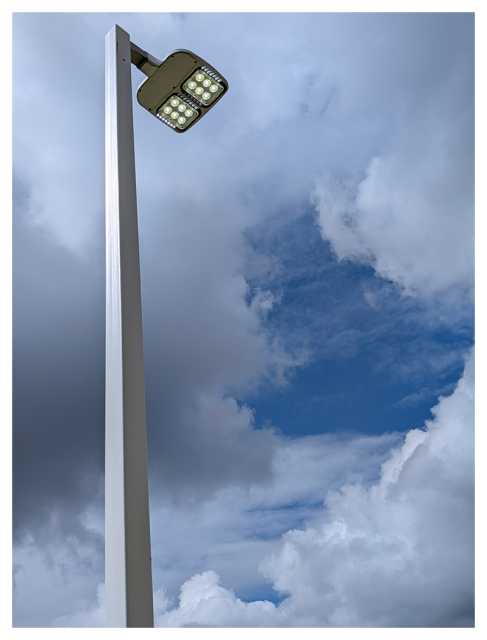 daytime as a storm approaches. a square, white pole rises at angle from bottom to top at left, a two-lamp LED light at the top. the background: large popcorn clouds curtain a dark blue sky with wispy clouds. 