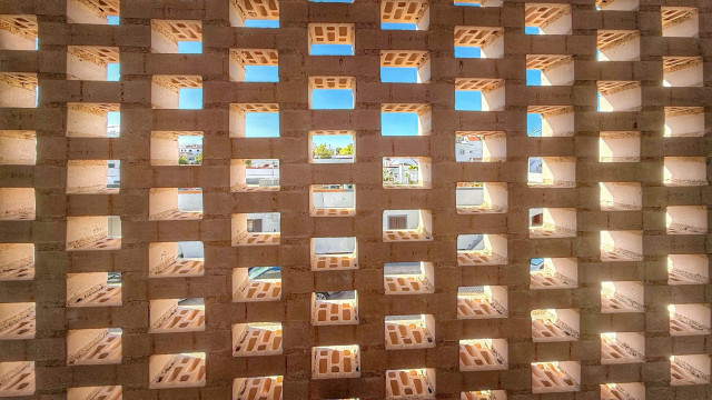 A textured wall made of light-colored bricks with square cutouts, allowing visible blue sky and distant buildings to be seen through the openings.