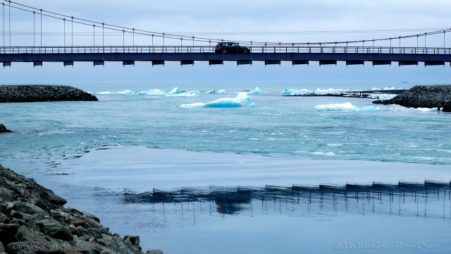 A photo of a river passing under a suspension bridge. There is a car about halfway across, and reinforcing blades hang beneath. The foreground is a manufactured rocky bank made of boulders, more can be seen in the distance containing the water flow. The water is a strong cyan blue, and there are icebergs floating in it. In the foreground is also the reflection of the bridge and a triangular surface formation at the pinch point, angled towards the sea. A black beach can be seen in the distance, covered in grounded bergs.