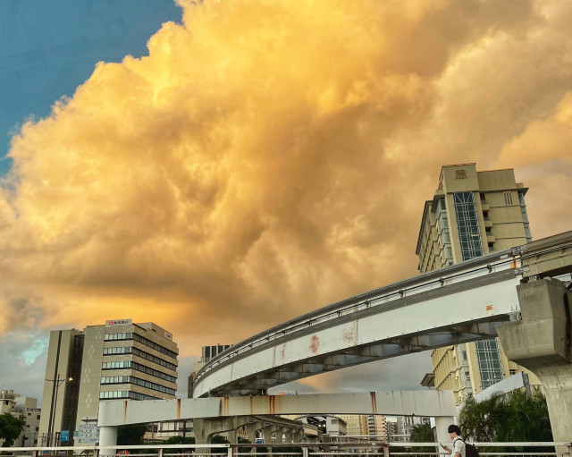 A vibrant sky filled with golden clouds contrasts against urban buildings and a monorail track. A person is seen walking below, adding a sense of scale to the scene.