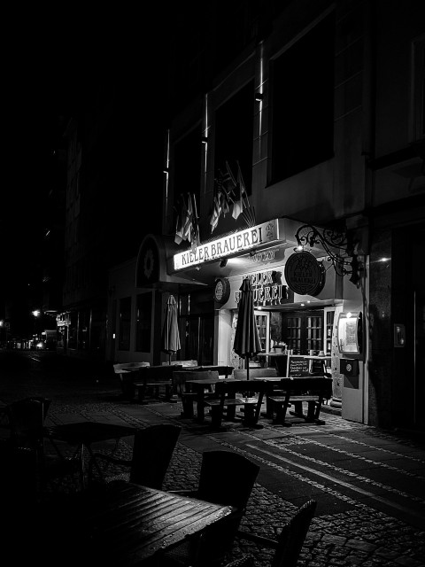 Das Foto zeigt eine nächtliche Straßenszene mit einem Fokus auf eine Einrichtung namens “KIELER BRAUEREI.” Das Bild ist in schwarz-weiß. Vor der Brauerei stehen Tische und Stühle im Freien. Die Beleuchtung kommt aus dem Inneren des Gebäudes und von einigen Straßenlaternen, die die Umgebung erhellen. Es sind keine Menschen auf dem Bild zu sehen, da es zu einer späten Stunde aufgenommen wurde. 

The photo shows a nocturnal street scene with a focus on a facility called “KIELER BRAUEREI.” The image is in black and white. There are tables and chairs outside in front of the brewery. The lighting comes from the interior of the building and from some street lamps that illuminate the surroundings. There are no people in the picture, as it was taken at a late hour.