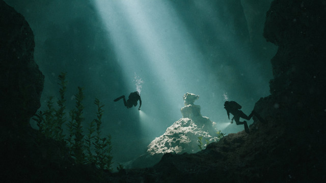 3D render of two divers exploring a cave. Light rays break through the turquoise water, illuminating a forgotten stone statue on the ocean floor. The statue is actually Suzanne, the default monkey head from Blender.