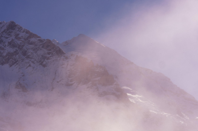 A majestic snow-covered mountain peak shrouded in misty purple and pink hues. The rugged, rocky face of the mountain is partially visible through the ethereal fog, creating a dreamlike atmosphere. The sky transitions from a deeper blue at the top to softer lavender tones near the mountain, giving the scene a serene and otherworldly quality.