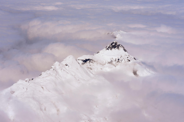 A dramatic view of snow-covered mountain peaks emerging from a sea of clouds. The central peak, jagged and majestic, rises prominently above the others, its rocky face partially visible beneath the snow. Wispy clouds swirl around the mountains, creating a dreamlike atmosphere. The sky has a soft, pastel quality with hints of blue and pink, giving the scene an ethereal and serene appearance.