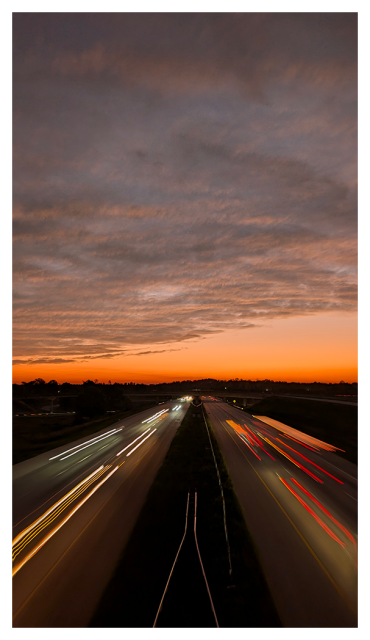 sunset. high angle viewfrom an overpass over four lanes of freeway with grass median that disappears into a treeline. the horizon behind the trees is dark orange and into pale blue gray with horizontal bands of gray/white/pink clouds.