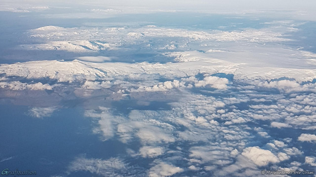 A photo of a landscape taken from a plane. Looking north, a range of mountains is visible. The sky below is cloudy but the coastline is just visible in the foreground. The peaks are iced over, on the left of the shot are three prominent ridges running horizontally across to the centre. On the right is a large, flat-topped mountain with steep sides, and beyond that is another line of crests.