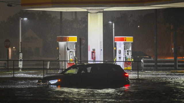 A photo of a car submerged in about two feet of water with a flooded gas station in the background. The brand colours of Shell are on the pumps and the canopy overhead but the photo is cropped to hide the Shell logo.