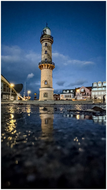 A lighthouse stands prominently against a twilight sky, reflecting in a wet surface in the foreground. Nearby buildings and streetlights create a serene ambiance.