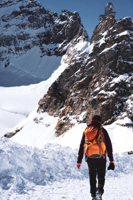 A hiker with an orange backpack walks through snow towards towering, snow-capped rocky mountains. In the distance, atop one of the highest peaks, sits a small observatory or scientific station with a distinctive dome. The rugged landscape is a mix of exposed rock faces and snow-covered slopes under a clear blue sky. Other visitors can be seen in the far distance, highlighting the scale of the mountainous terrain.