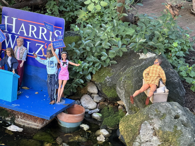 Display of Barbies on HarrisWalz campaign stage with JD Vance looking on from the side while fucking a couch with no pants on. 