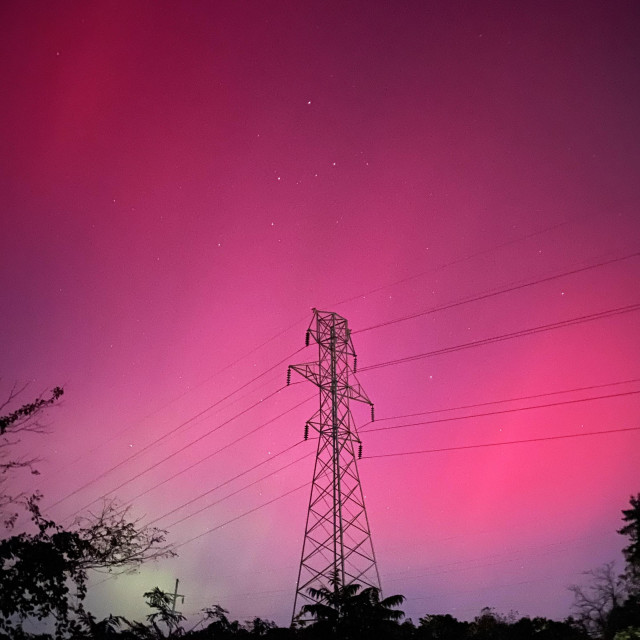 The sky above a transmission tower is aglow with mostly red and some green on a clear, star filled night.