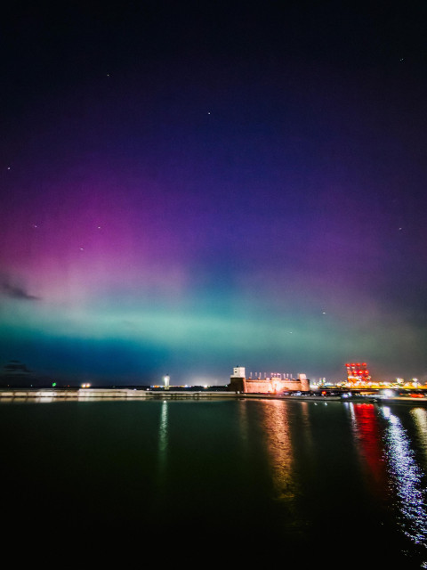 Purple and green aurora over a lake and fort. The car park is full and the lighthouse is just about visible.