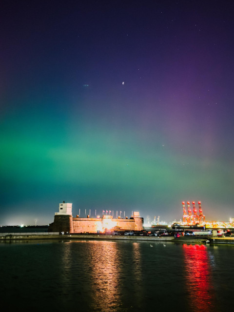 Purple and green aurora over a lake and fort. The car park is full and the lighthouse is just about visible.
There are red cranes in the distance and a couple of swans on the lake.