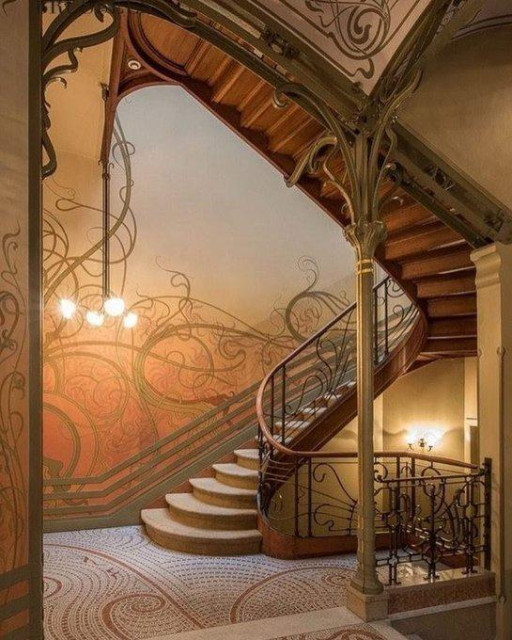 This image shows a beautifully ornate staircase in an Art Nouveau style building. The staircase has a curved shape with wrought iron railings and a wooden banister. The walls are decorated with colorful murals and mosaics, and there is a chandelier hanging from the ceiling.