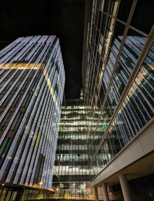 Wide-angle night photo of three large, glass-coated buildings, taken from below. At the rear, framed by the other two, is a block that is all horizontal green-yellow strips of lit office-space. On the left is one with just one floor lit from within, but vertical stripes of slim white lights cover every surface. On the right the building extends past the camera to exit shot at the top and right and mostly just reflects the other two.