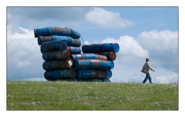 Kunstobjekt Stuhl aus blauen Ölfässern ca. 8 Meter groß auf einer Wiese. Rechts entfernt sich ein junger Mann. An object of art: a chair made of blue oil barrels, about 8 meters tall, in a meadow. A young man is walking away on the right.