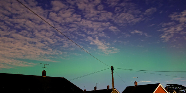 A band of light green spread out across the sky just above some rooftops. The rest of the sky is blue, with a light covering of cloud. Shot from the middle of a suburban town near London at 2023 GMT on 10 October 2024.