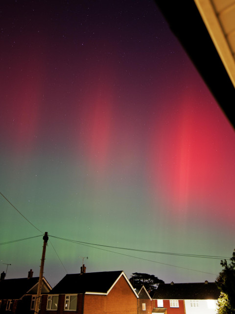 More defined streaks of green just above the rooftops in the frame, with the streaks turning bright red as they get higher into the sky. Shot from the middle of a suburban town near London at 2055 GMT on 10 October 2024.