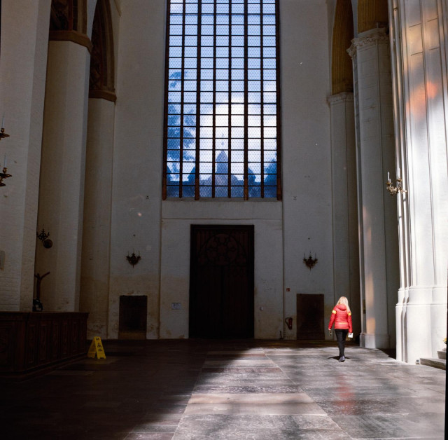 Das Foto zeigt einen Teil eines riesigen Fensters aus einer Kirchen in Stralsund. Die Sonne scheint hindurch und zeichnet die Rahmen der einzelnen Fensterelemente auf den Boden der Kirche.
Unten rechts ist eine Frau zu sehen, sie wirkt eher winzig in dem Gang der Kirche. 