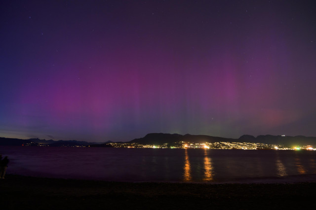 a long purple arc aurora frames the mountains near the horizon, as the water of the harbour reflect the faint purple glow as well as the city lights on the opposite shore