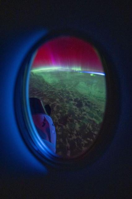 Looking out through a porthole-style window inside the Dragon Endeavor capsule, this photography shoes red, pink, and green aurore above the cloudy atmosphere of Earth. The porthole is lit with blue light, and another Dragon capsule can be seen in the bottom left of the window.