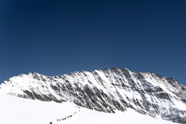 A panoramic view of a snow-covered mountain range against a deep blue, cloudless sky. The jagged peaks are brilliantly lit, creating a stark contrast between the bright snow and dark rock faces. In the foreground, a long line of tiny figures, presumably hikers or climbers, can be seen trekking across a snow field towards the mountains. The scale of the landscape dwarfs the human presence, emphasizing the grandeur and challenge of the alpine environment. The image captures the allure of mountaineering and the raw beauty of high-altitude landscapes.