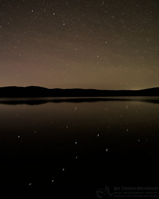 A landscape photo taken during deep twilight of the seven star asterism know as the Big Dipper, which looks like a large ladle. Three stars make up its handle and four stars make the dipping cup at the end. While it is hard to see among all of the stars in the sky it is easily seen reflected in the water of a large lake surrounded by mountains.