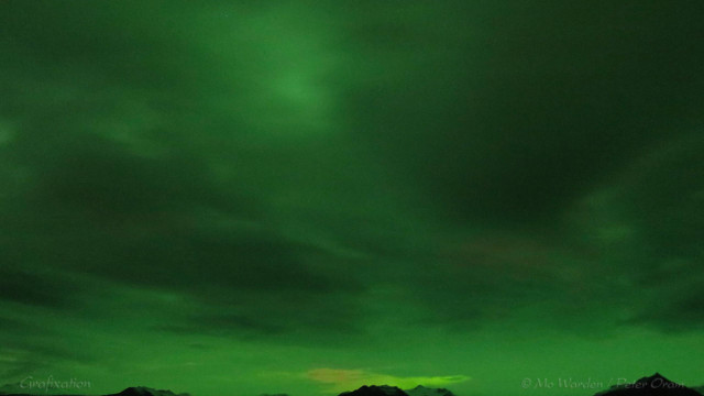 A photo of the night sky, almost completely full of dark cloud. At the bottom of the shot is a narrow line of mountain tops, and a tiny window in the cloud which is showing some colour behind it, reds and greens. The rest of the clouds are heavily tinted with a lurid dark green, in places almost completely obscured and in others slight visibility.