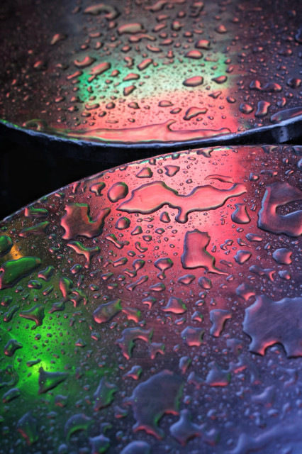 Photograph shows raindrops gathered on metallic table tops in which nearby red and green restaurant lights are colouring the scene like an aurora.