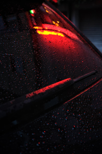 The bright lights of a restaurant are reflected in the side window of a parked car on which tiny raindrops are shining like coloured gems.