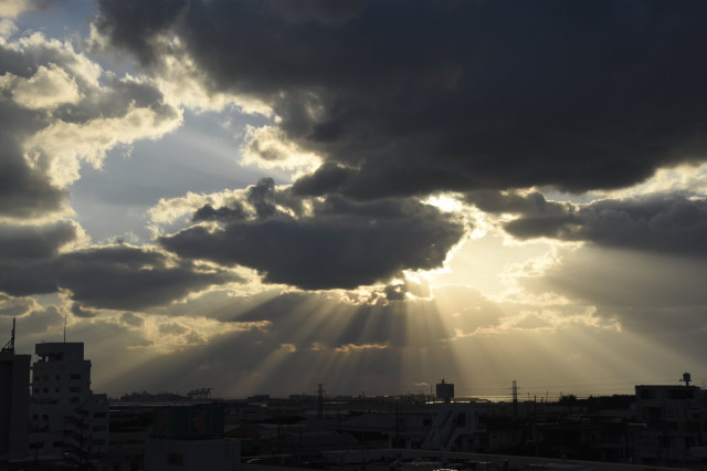 A dramatic sky filled with clouds, illuminated by rays of sunlight breaking through. The foreground features silhouettes of buildings and structures, creating a contrast against the bright background.