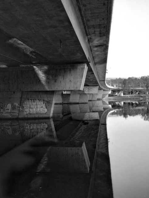 Black And White image of a bridge from the banks of the river. The reflection in the water of the bridge is like a mirror