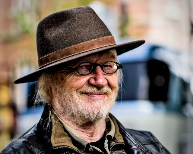 Portrait eines älteren Herren mit weißen Haaren und Bart. Er trägt einen eleganten Filzhut und eine kleine, runde Brille. Der Herr lächelt entspannt und gut gelaunt in die Kamera. 
.
Portrait of an elderly gentleman with white hair and beard. He is wearing an elegant felt hat and small, round glasses. The gentleman smiles relaxed and in a good mood into the camera. 
.
