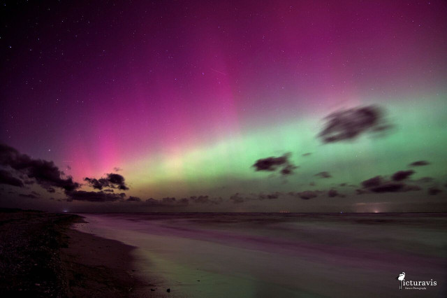 A photograph of the northern lights over a coastal landscape. The aurora spans the entire image, with the upper parts in vivid pink and the lower parts in yellow and green. The colors are reflected off the long exposure smoothed seawater in the lower part of the image. Beach sand is visible in the lower right corner, with many larger rocks. Some clouds are moving across the sky. A few lights in red, blue and white are visible on the horizon.