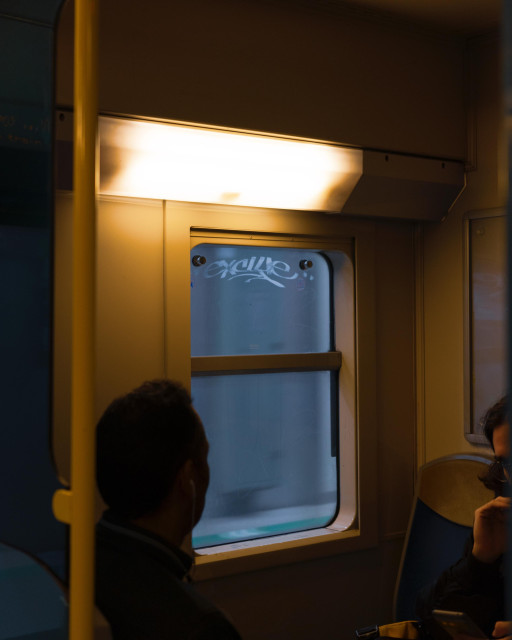 Interior view of a train or subway car at night. The image is dominated by warm, golden lighting from overhead fixtures. A large window takes up the center of the frame, its glass etched with graffiti. The silhouettes of two passengers are visible in the foreground, one on each side of the image. The atmosphere is quiet and introspective, capturing a moment of urban transit during off-peak hours. The contrast between the bright interior and the dark exterior visible through the window creates a sense of cocoon-like safety within the public transportation space.