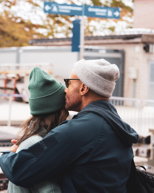 A couple embraces at a train station. The man, wearing a gray beanie and dark jacket, kisses the forehead of a woman in a green beanie. They stand on a platform with directional signs visible in the blurred background, creating an intimate moment amidst the public setting.