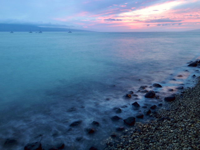 A long exposure of a serene ocean scene at dusk, featuring a calm sea with gentle waves lapping against a rocky shore. Several boats are anchored in the distance under a pastel-colored sky transitioning from pink to blue. Photo was taken in 2019, standing in old town Lahaina looking out over the ocean.