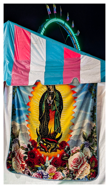 night at the country fair. a blanket with elaborate art featuring Mary hangs on the side of a white vendor's tent, under a triangular flap of blue/white/pink vinyl  awning. the background is a lighted ride, a half-circle with flags on top. the sky is black.