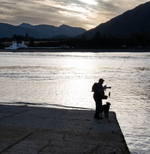 An angler holds a recently caught fish on his rod. His curious dog looks on. The pair are in silhouette as light from the setting sun bounces on the water behind them. 