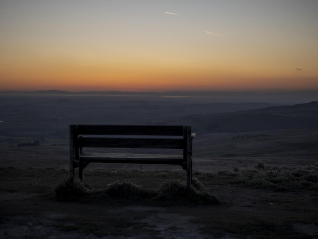 A wooden bench stands at the summit of a mountain pass with a view across the Cumbrian countryside. The Cumbrian coast can just be made out in the distance. The photo was taken just after sunset so the horizon has a deep range colour. 