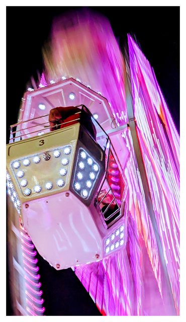 low angle view of a Ferris wheel-type ride. a car in the foreground carries one person and is in focus. the rest of the ride is a motion-blurred in a dramatic way. mostly tones of pink with touches of pale gold against a black night sky.