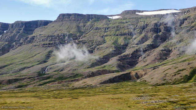 A photo of a mountainside with a mostly clear cyan sky above. The foreground is green pasture and is scattered with the white dots of grazing sheep. A glacial mountain side fills most of the shot, steep-sided in the upper regions and curving gently down to flatten out at ground level. Water has gouged valleys into this rock, and most of them seem to contain a waterfall. Some of the upper reaches hold ice patches in the sheltered spots, and scraps of wispy cloud are clinging to the lower slopes.