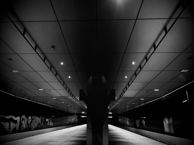 Black and white photo of an emty platform in a subway station with tracks on either side of the frame. In the middle is a large support structure. On the walls are various decorations of objects.