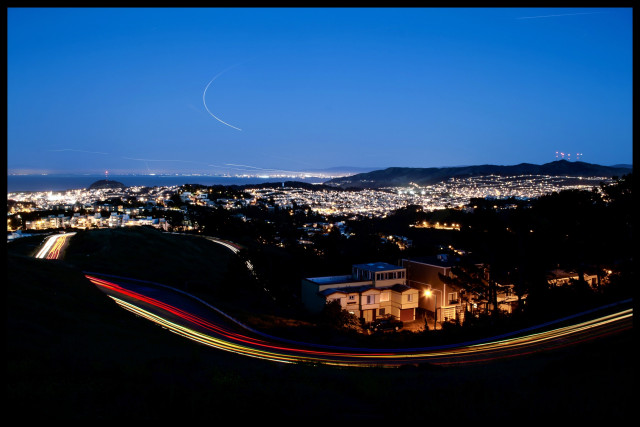 A blue-hour view looking southeast from an elevation of about 900 feet, across a residential section of San Francisco, the Bay, and a bit of the East Bay in the distance.

Royal blue skies, white and amber city lights, and a few light trails of planes taking off from Oakland Airport. Some light trails from cars swooping past on the road leading up to the Peaks. And lots of dark areas in the foreground where shrubbery sits quietly, doing nothing.