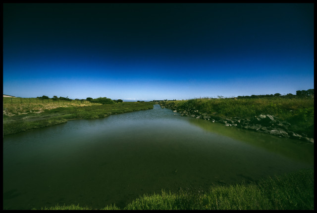 An inlet of water at Heron's Head Park. The inlet leads out to the San Francisco Bay on the horizon.

On the banks of the inlet: overgrown grass and a bit of rock.

Above: weird sky with a gradient that transitions from black above to royal blue to almost white across the bay. 

It's peaceful. I don't like it.