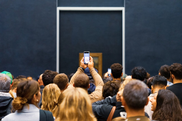 A crowded room at the Louvre, with a sea of visitors' heads in the foreground. In the center, a person's arms are raised high, holding a smartphone to take a photo of what is presumably the Mona Lisa. The painting itself is not visible, obscured by the crowd and distance. The background shows a large, dark wall with a framed empty space, highlighting the irony of the scene - the real spectacle is the mass of people rather than the artwork they've come to see.