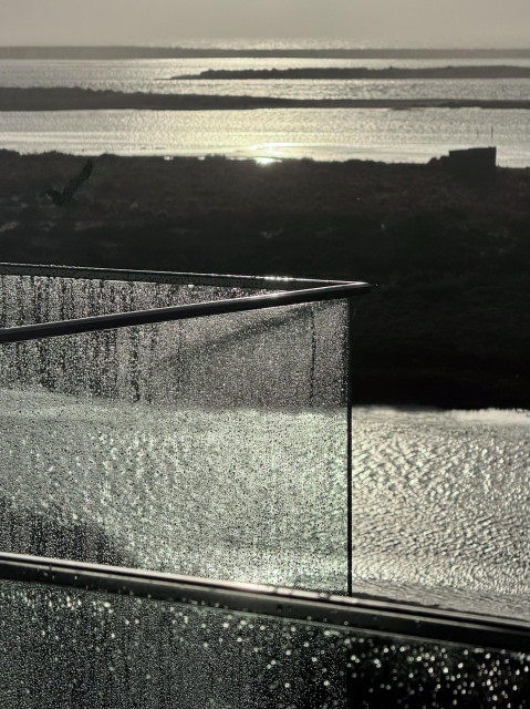 A close-up of a wet glass railing, with droplets reflecting light. In the background, a shimmering body of water is visible, along with distant landforms under a hazy sky. A bird can be seen in the upper left corner.